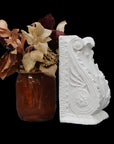 photo of white plaster cast sculpture of wall bracket with scrolls and acanthus leaves placed on a surface next to a red vase of red and off-white flowers against a black background