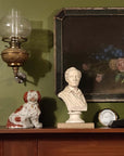 photo of off-white plaster cast sculpture bust of man, namely the poet Lord Byron, on socle base atop book on mantel beside foo dog with painting and antique globe light on wall