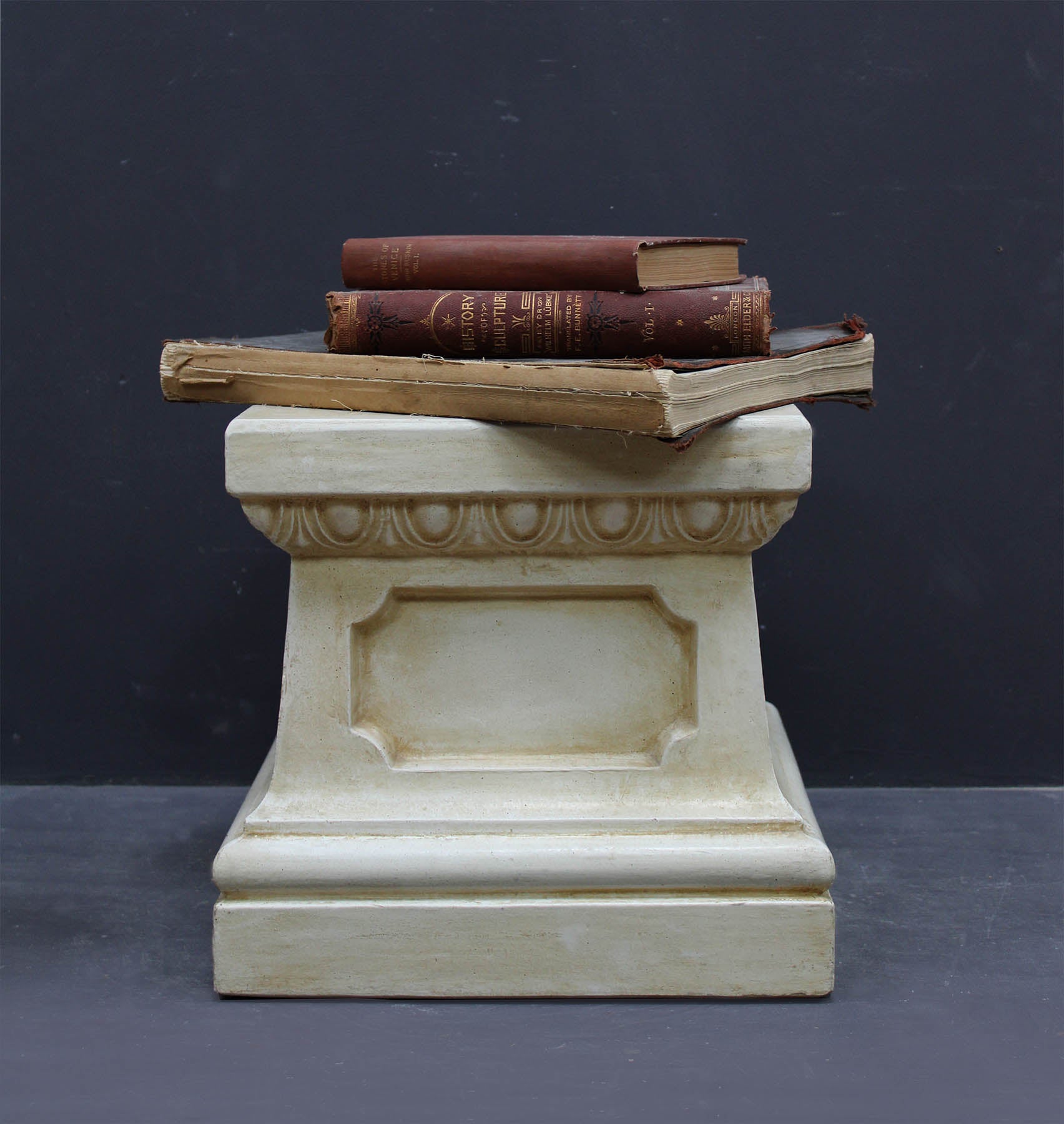 photo of off-white plaster cast reproduction of short, rectangular pedestal with three red books stacked on top against a gray background