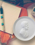 Photo of a white plaster cast of a medallion with a profile of a man wearing a wrath on his head, on a surface next to an open blank diary, pen, camera and orange patterned scarf.