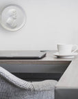 Photo of a white plaster cast of a medallion with a profile of a man wearing a wrath on his head, on a wall behind a desk with a vase, computer, coffee cup, and picture frame.