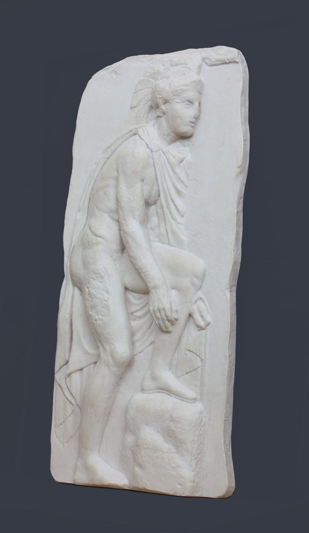 photo of off-white plaster cast relief sculpture of male nude with cloak bending to tie his sandal against gray background