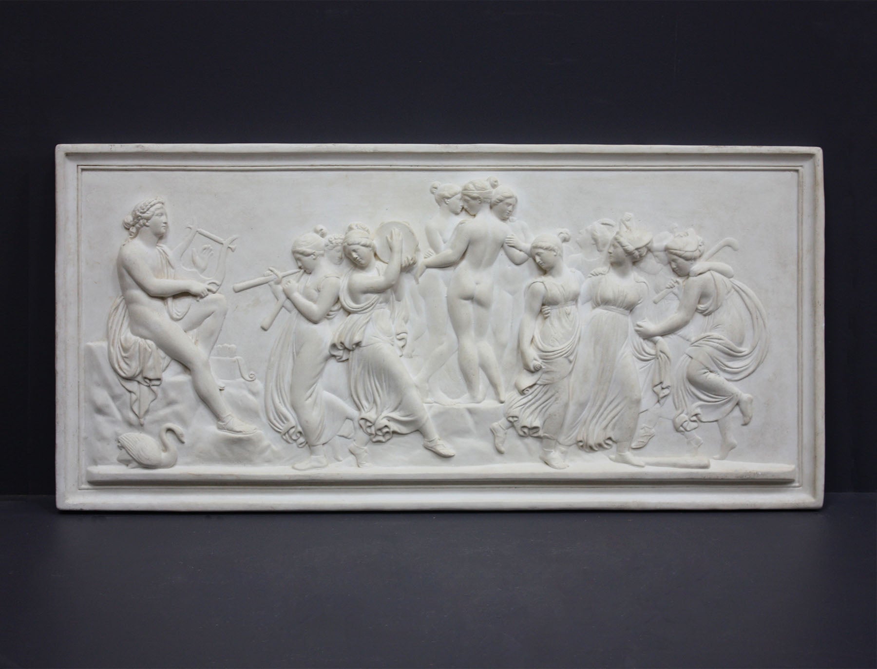 Photo of plaster cast sculpture relief of nude and clothed females (muses) dancing and playing instruments and one male (Apollo) playing a lyre on a dark gray background