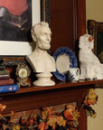 plaster cast bust sculpture of man with beard, namely Lincoln, on dark wood mantel with blue plate, white foo dog, and bronze clock and painting behind