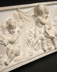 side view photo of white plaster cast with warm tones of relief sculpture of several cupids talking together with books and baskets of fruit on a black background