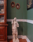 photo of plaster cast of female figure, namely the goddess Diana, with robes on a wood floor in front of a green wall with part of a hutch visible to the left and decorations in hutch and on walls