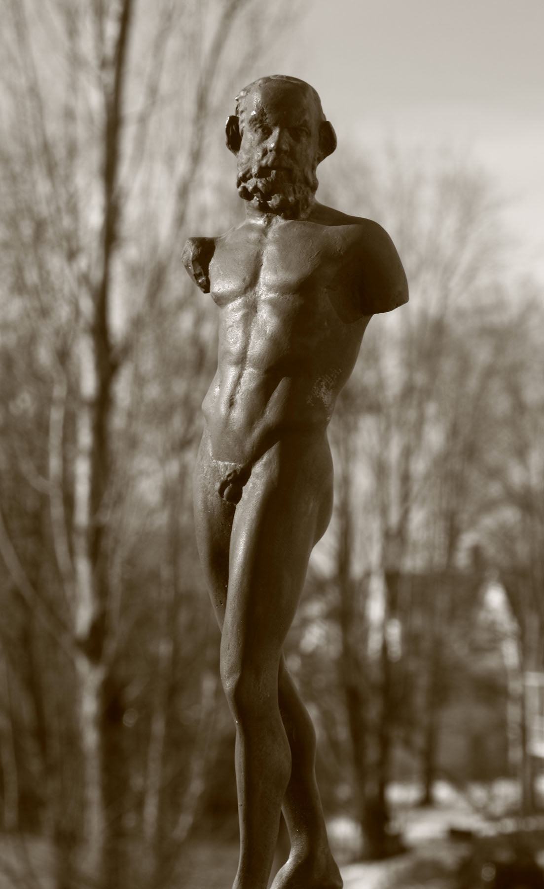 photo detail of bronze-colored plaster cast sculpture of standing nude satyr figure on square base in front of trees