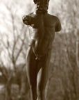 photo detail of bronze-colored plaster cast sculpture of standing nude satyr figure on square base in front of trees