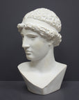 photo of white plaster cast sculpture bust of female, namely the goddess Athena, with short, curly hair and headband on gray background