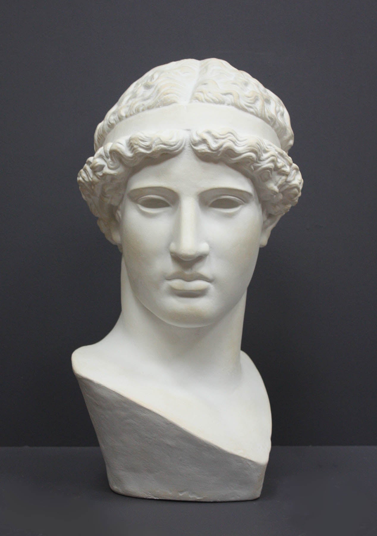 photo of white plaster cast sculpture bust of female, namely the goddess Athena, with short, curly hair and headband on gray background