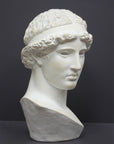 photo of white plaster cast sculpture bust of female, namely the goddess Athena, with short, curly hair and headband on gray background