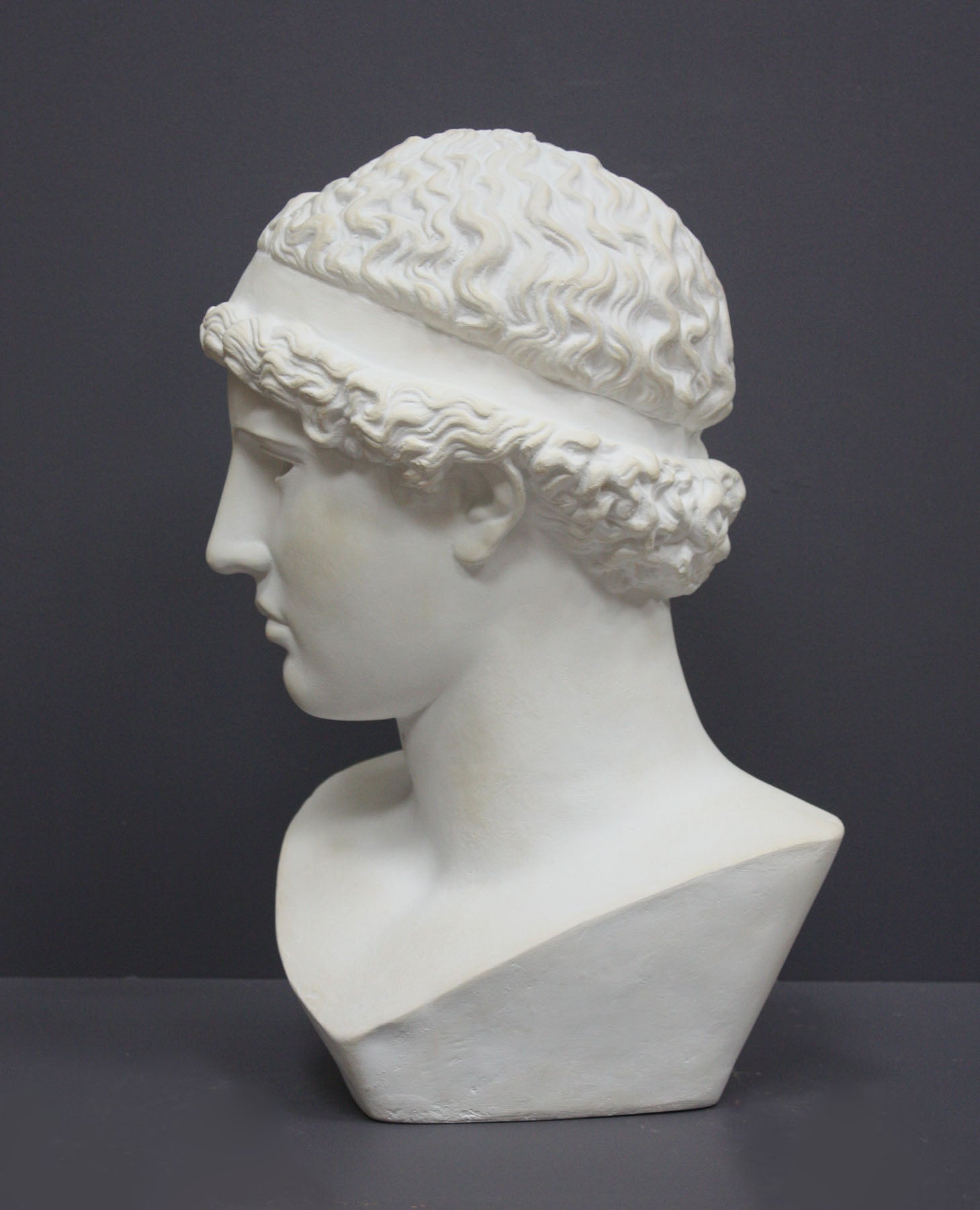 photo of white plaster cast sculpture bust of female, namely the goddess Athena, with short, curly hair and headband on gray background