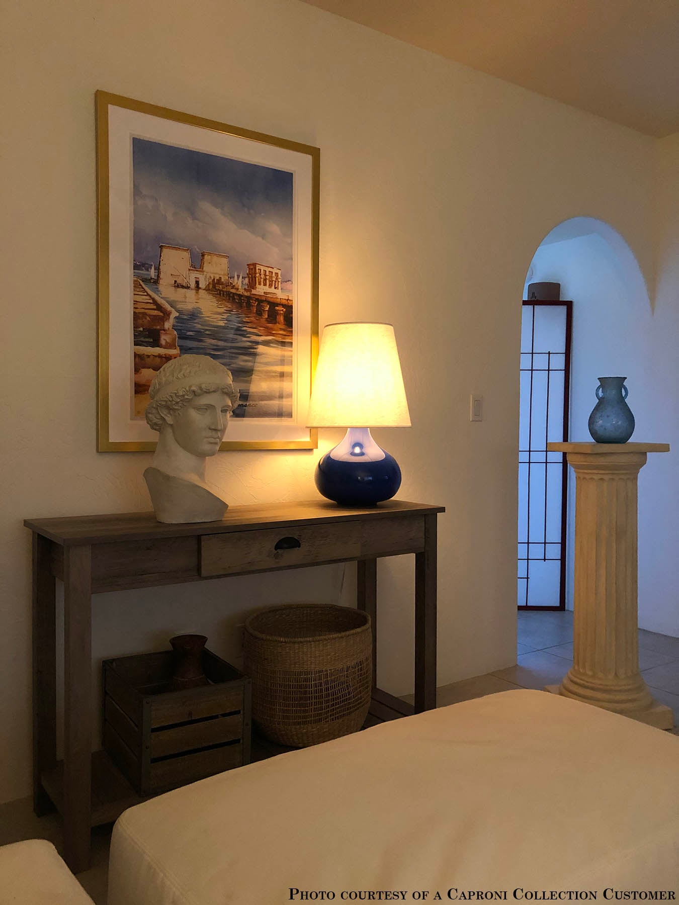 plaster cast sculpture bust of female figure, namely the goddess Athena, with short, curly hair and headband on a wooden table next to blue lamp and in front of painting in white-walled room