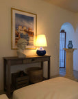 plaster cast sculpture bust of female figure, namely the goddess Athena, with short, curly hair and headband on a wooden table next to blue lamp and in front of painting in white-walled room