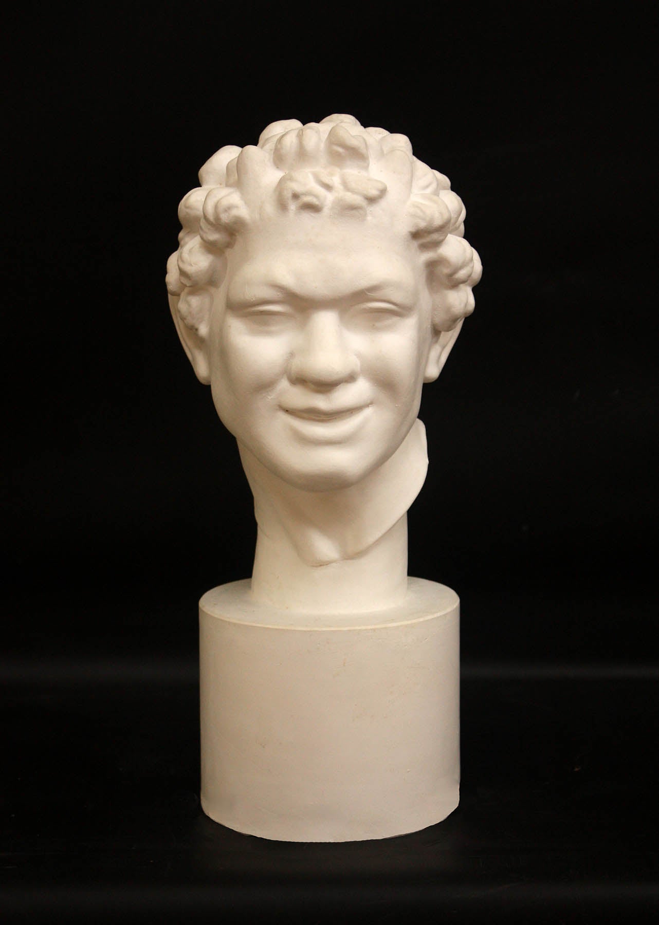 Photo of white plaster cast sculpture of faun head on base against black background