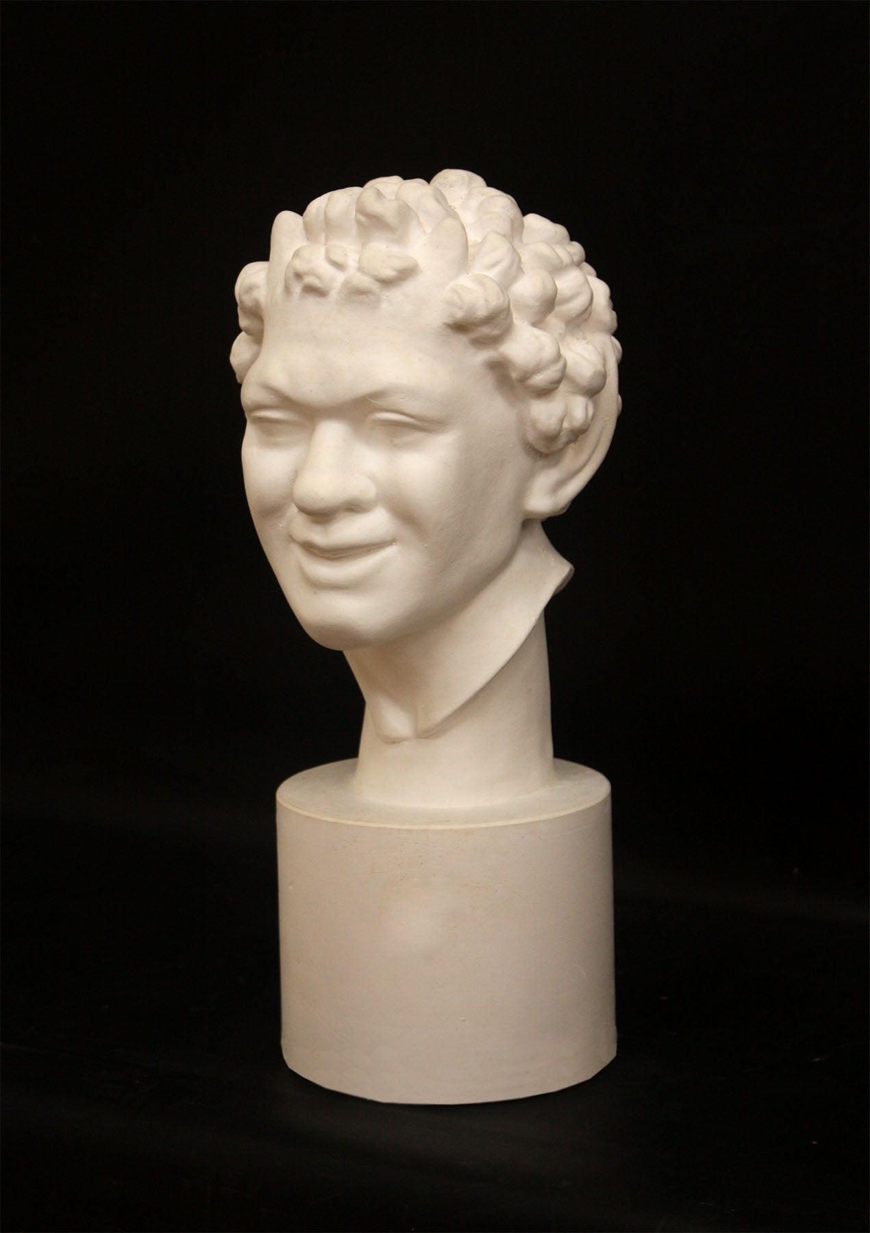 Photo of white plaster cast sculpture of faun head on base against black background