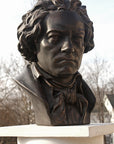 photo of bronze-colored plaster cast sculpture bust of man, namely Beethoven, with neckerchief on pedestal with sky and trees in background