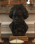 photo of bronze-colored plaster cast sculpture bust of man, namely Beethoven, with neckerchief on dark wood table with candlesticks