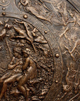 detail photo of bronze-colored plaster reproduction of a shield with intricate relief sculpture set against black background