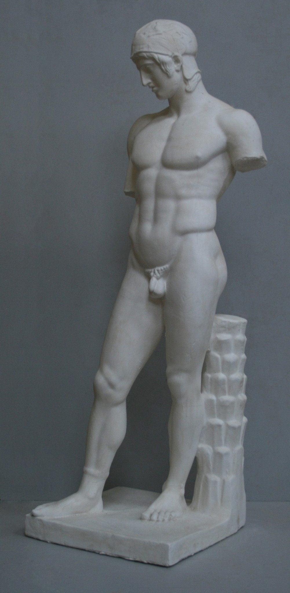 photo of plaster cast of sculpture of nude male, namely the god Ares, without arms and wearing a helmet with a supporting tree trunk, on a gray background