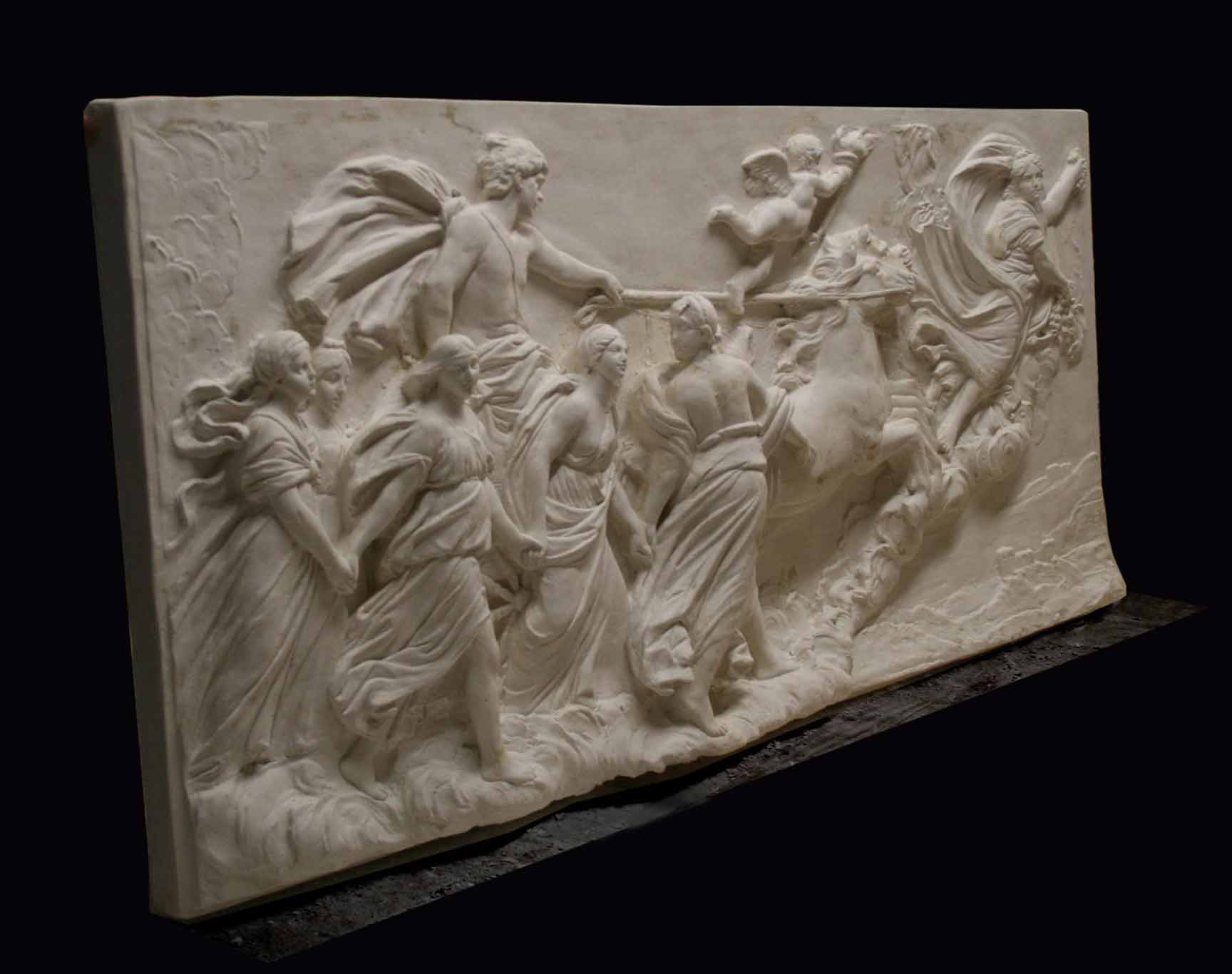 photo of a plaster cast sculpture relief of a woman, namely the goddess Aurora, flying and leading a chariot with a man pulled by horses towards the right while a cherub and other women fill the scene, on a black background