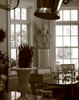 Sepia-toned photo of male bust sculpture on tall pedestal in between two windows in a dining room