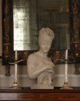 photo of plaster cast sculpture bust of Madame Recamier with high up-do and ribbon over forehead and hands crossed over chest covered in cloth on a dark wood table with bronze decorations, candlesticks, and bronze framed mirror and two pictures