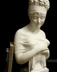 photo with black background of plaster cast sculpture bust of Madame Recamier with high up-do and ribbon over forehead and hands crossed over chest covered in cloth, and two books leaning against her