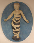 photo of plaster cast relief of child with mummy-like wrappings on a blue oval background on a tan-colored wall