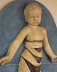 closeup photo of plaster cast relief of child with mummy-like wrappings on a blue oval background on a tan-colored wall