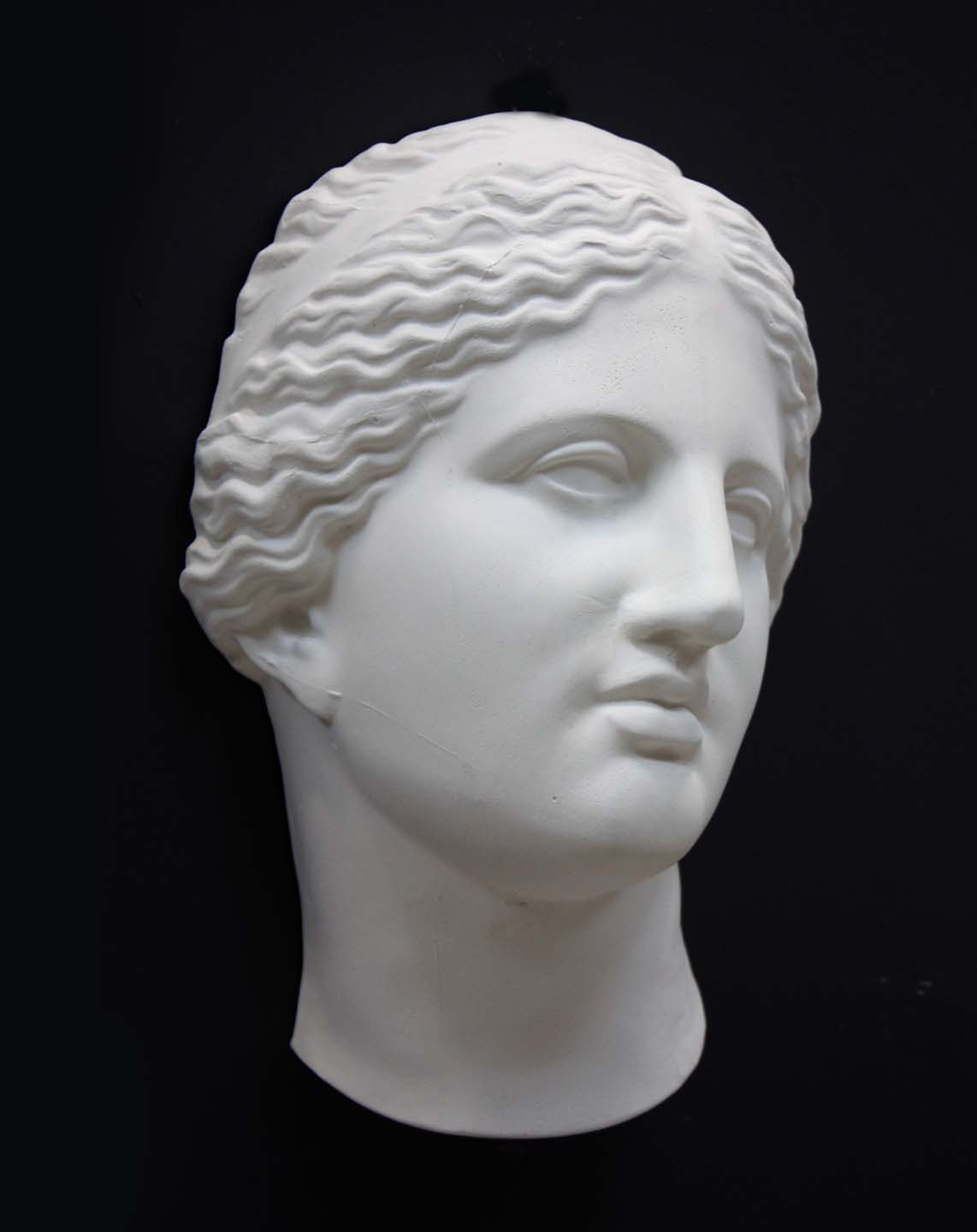 photo of white plaster cast of sculpture of face of Aphrodite of Knidos with wavy, pulled-back hair on dark gray background