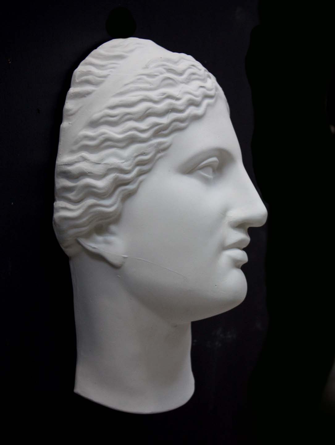 photo of white plaster cast of sculpture of face of Aphrodite of Knidos with wavy, pulled-back hair on dark gray background