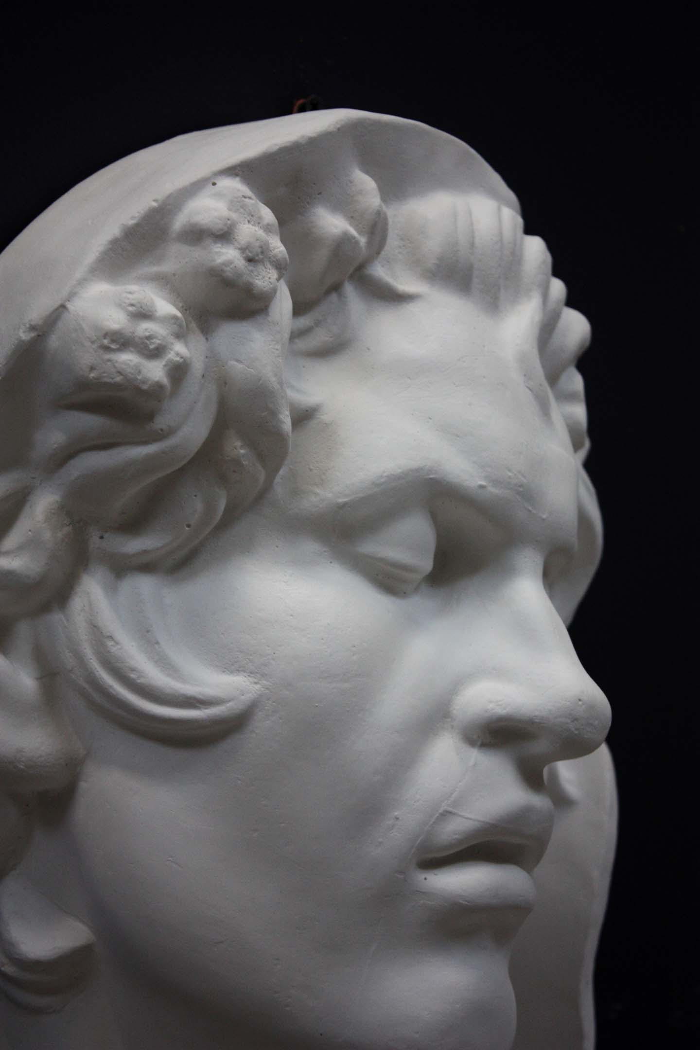 photo of plaster cast of sculpture of face of Sleeping Faun with two flowers in curly hair on dark gray background