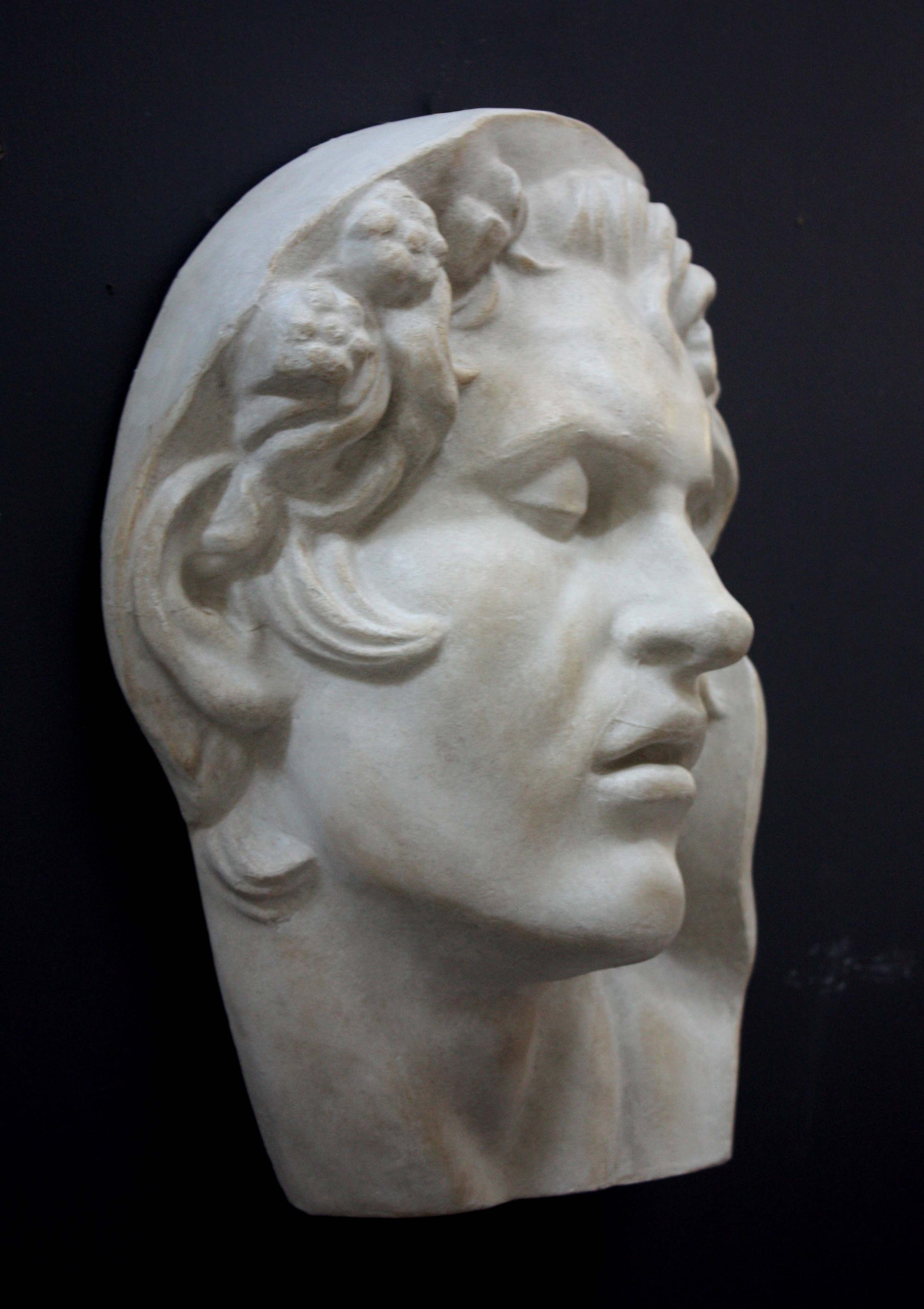 photo of plaster cast of sculpture of face of Sleeping Faun with two flowers in curly hair on dark gray background