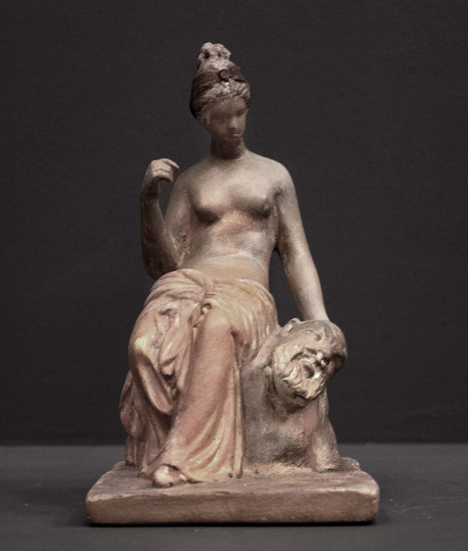 photo of plaster cast sculpture of female half nude with pink robes seated and holding a mask, set against gray background