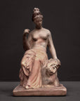 photo of plaster cast sculpture of female half nude with pink robes seated and holding a mask, set against gray background