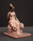 photo of plaster cast sculpture of female half nude with pink robes seated and holding a mask, set against gray background