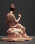 photo of plaster cast sculpture of female half nude with pink robes seated and holding a mask, set against gray background