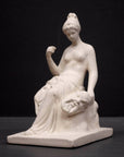 photo of white plaster cast sculpture of female half nude seated and holding a mask, set against gray background