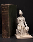 photo of white plaster cast sculpture of female half nude seated and holding a mask, set beside green book and against gray background