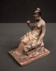 photo of plaster cast sculpture of female half nude with pink robes seated and holding a mask, set against gray background