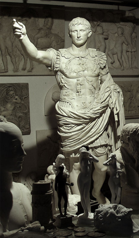 photo of white plaster cast sculpture of a man with armor and robes standing in contrapposto with his right arm raised, namely Augustus Caesar, in a gallery with relief sculptures hanging behind and small 3D works on table in foreground