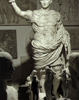 photo of white plaster cast sculpture of a man with armor and robes standing in contrapposto with his right arm raised, namely Augustus Caesar, in a gallery with relief sculptures hanging behind and small 3D works on table in foreground