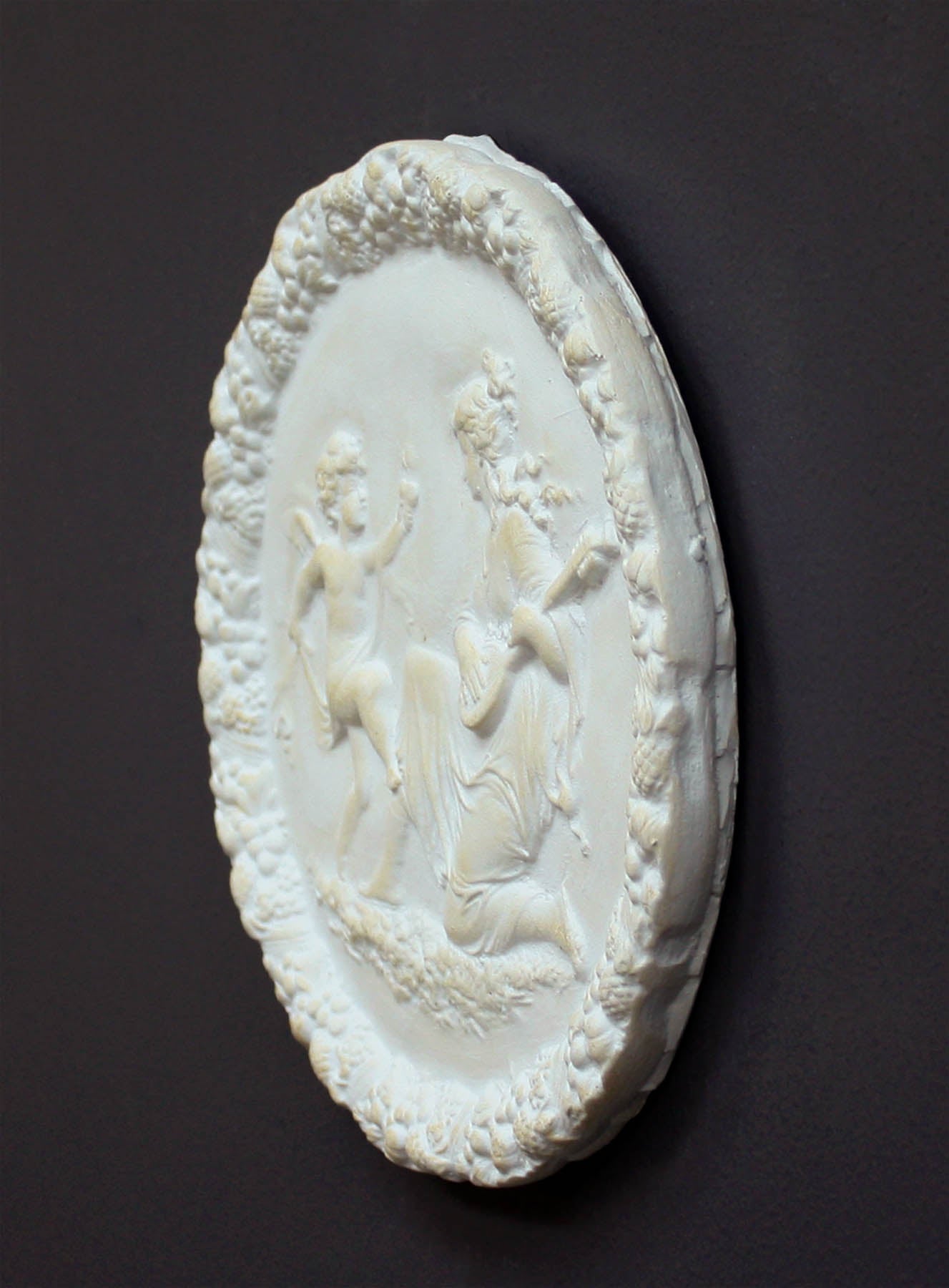 photo of white plaster cast relief sculpture of a child dancing and a female figure playing an instrument against dark gray background