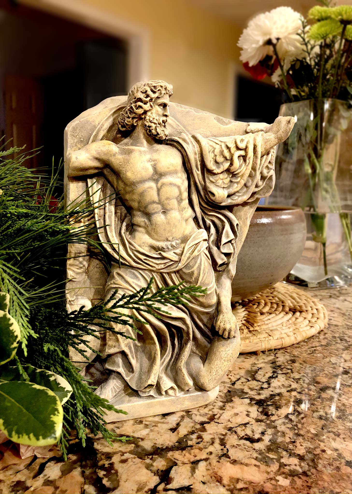 photo of plaster cast relief fragment of man, namely the god Zeus, on a red-orange granite counter leaning against a stone bowl with a plant visible on its left and a vase of flowers on its right