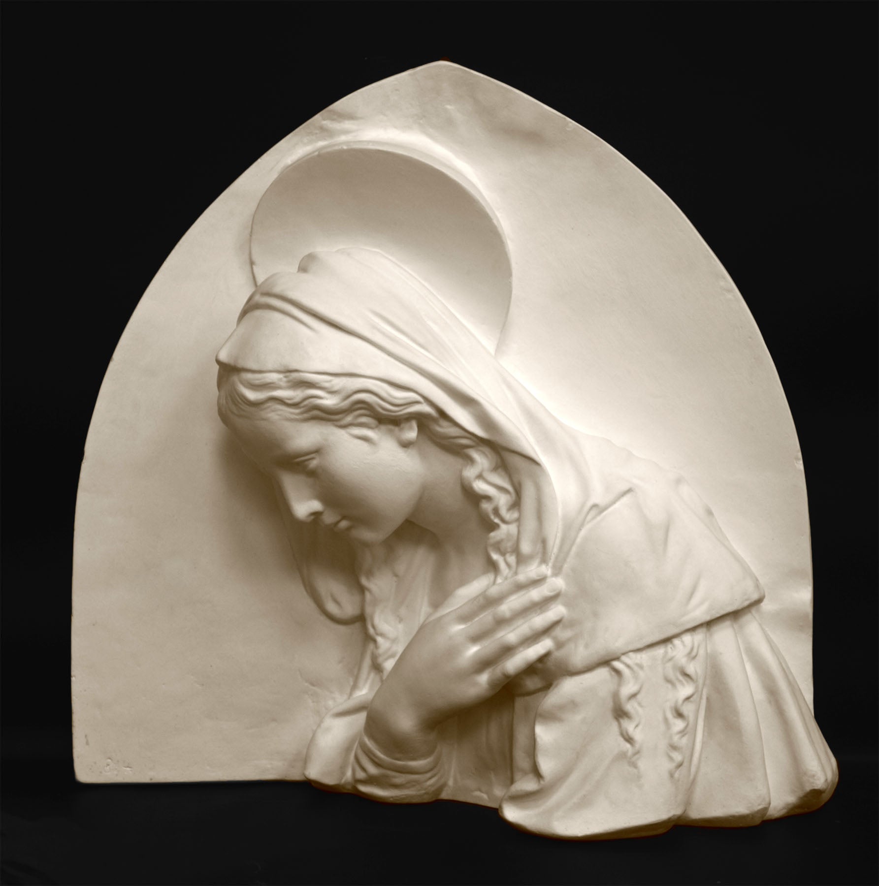 photo of plaster cast sculpture relief of the Madonna leaning over with her right hand crossed over her chest and a halo above her head on a black background