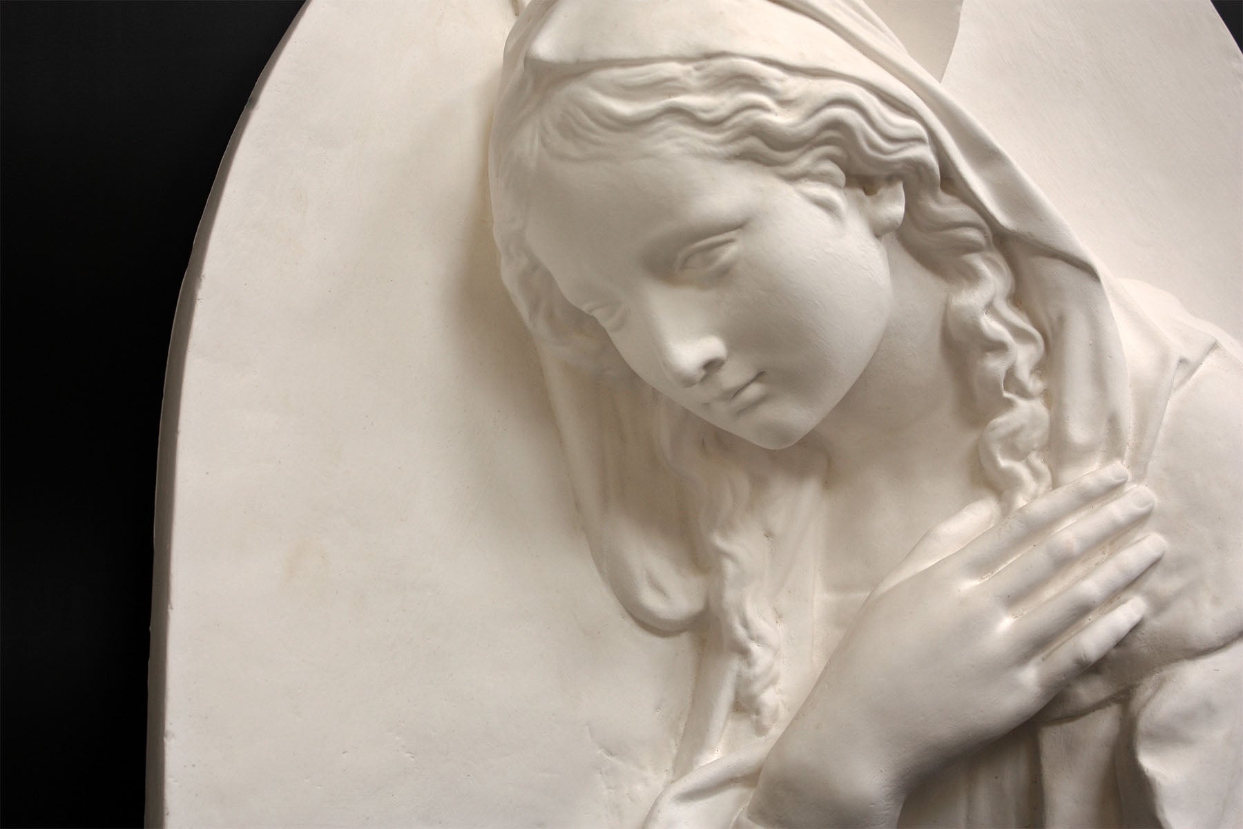 photo of plaster cast sculpture relief of the Madonna leaning over with her right hand crossed over her chest and a halo above her head on a black background