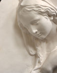 photo of plaster cast sculpture relief of the Madonna leaning over with her right hand crossed over her chest and a halo above her head on a black background