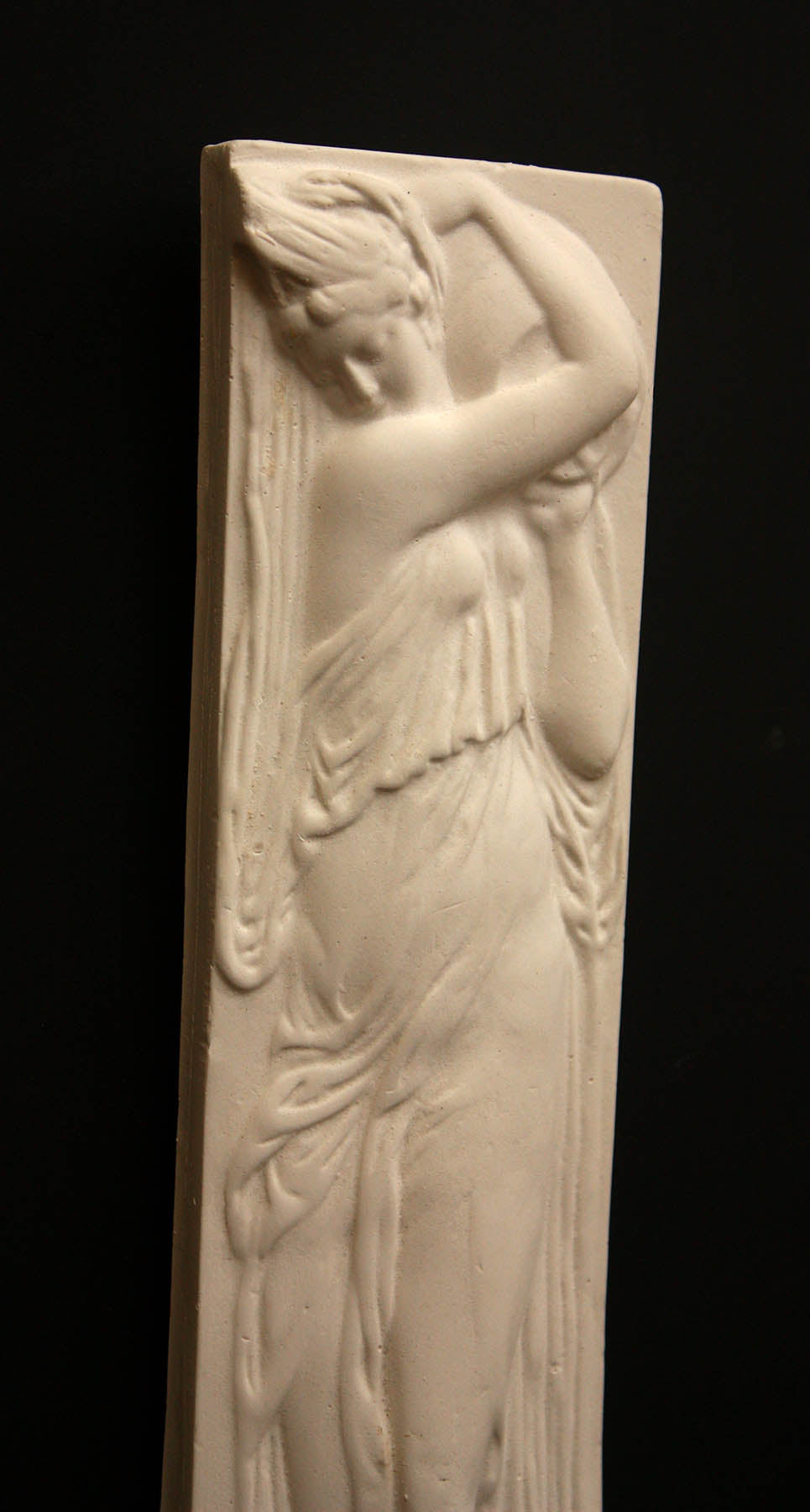 photo of plaster cast relief sculpture of female in drapery carrying jug over shoulder against black background