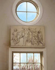 Photo of a plaster cast of figures and an element on a tan wall above a door and below a round window with blue sky coming through